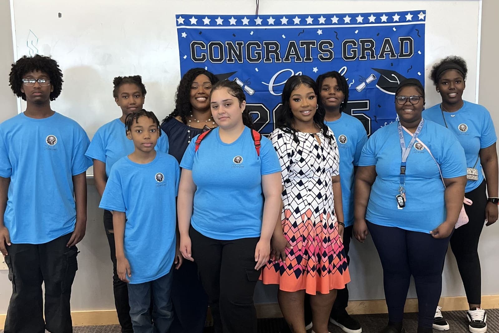 Nine young Brilliantaires volunteers in blue volunteer shirts standing with a staff member in front of a 2025 graduation banner
