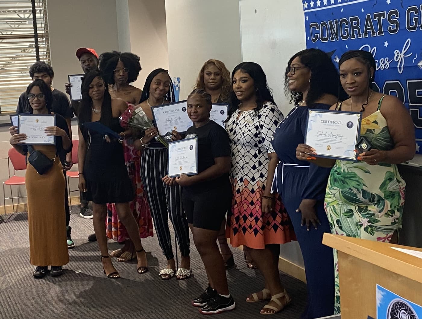 A Brilliantaires class standing together after graduation, most of them holding their certificates, in front of a Congrats Grad banner