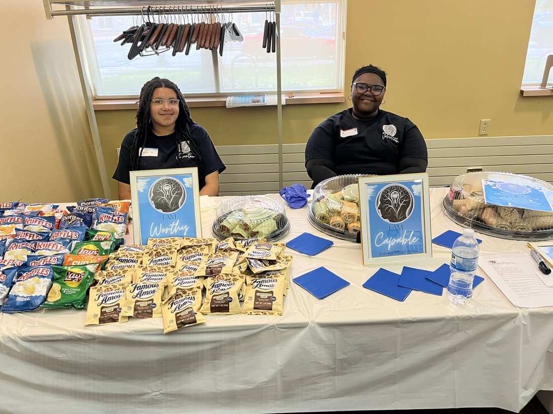 Two Brilliantaires staff in branded polo shirts behind a welcome table laid out with food, beside framed signs reading Worthy and Capable